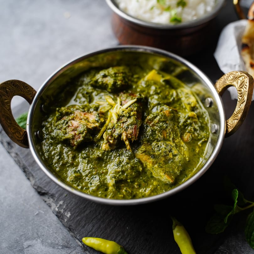 A Mahek Indian spinach curry with chicken in a brass-handled bowl, with rice and naan in the background.