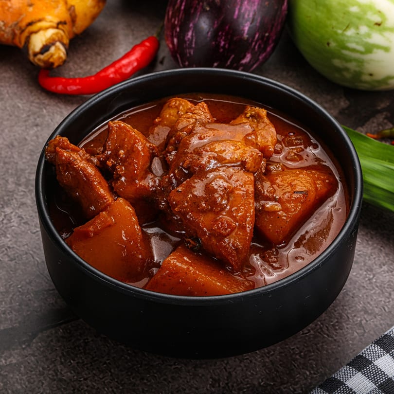 Close-up of Mahek's Indian chicken curry in a black bowl, surrounded by fresh vegetables.